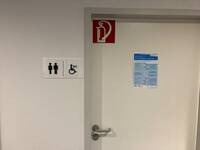 Ein helle Türe in einer weißen Wand. Links davon sind Symbole für Behindertentoilette, Herren und Damen an der Wand