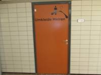 orangefarbene Türe in einer weiß gekachelten Wand, auf der Tür ist der Schriftzug UMKLEIDE so wie Symbole: Mann, Kleiderbügel