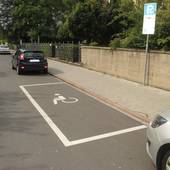 ein auf der Straße an der Gehwegseite eingezeichneter Behindertenparkplatz mit Rollstuhlsymbol auf dem Boden, anschließend daran eine Mauer, davor Parkschild