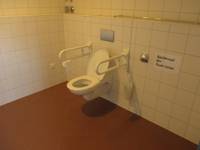 Toilette an einer weiß gekachelten Wand mit einem Haltegriff rechts und links. Rechts an der Wand ein Spülknopf mit einem großen Schild das auf den Knopf hinweist. Der Boden ist braun gefliest