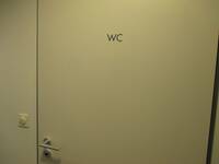 Eine helle Türe mit einem dunklen Rahmen in einer weißen Wand. Im oberen Drittel befindet sich mittig ein Schild mit der Aufschrift: WC