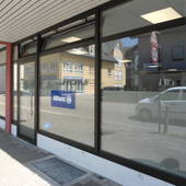Im EG mehrere große Schaufenster und Glastüren in einer Reihe. Im rechten Bildbereich Eingangstüre zum Büro. im