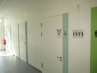 Eine weiße Tür in einer weißen Wand. Auf der Tür ist ein Symbol für alle Geschlechter und ein Rollstuhlsymbol. Rechts neben der Tür ist ein schmaler, senkrecht verlaufender hellblauer Streifen.
