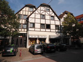 Fachwerkhaus mit Spitzdach. Im EG mit kleiner Markise überdachte Schaufenster, in der Mitte Eingangstür. Am Bildrand links und rechts Bäume.