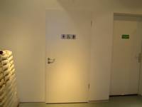 Auf dem Bild sieht man eine weiße Eingangstür mit Symbol der Behindertentoillette die Wand darum herum ist weiß 