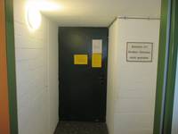 dunkle Tür in einem kurzen Flur, auf der Tür sind mehrere Zettel angebracht. Links von Tür befindet sich eine runde Wandleuchte