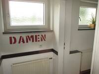 Eine offenstehende Türe in einer weißen Wand, unter dem Fenster vor der Tür steht in großen Buchstaben der Schriftzug: DAMEN